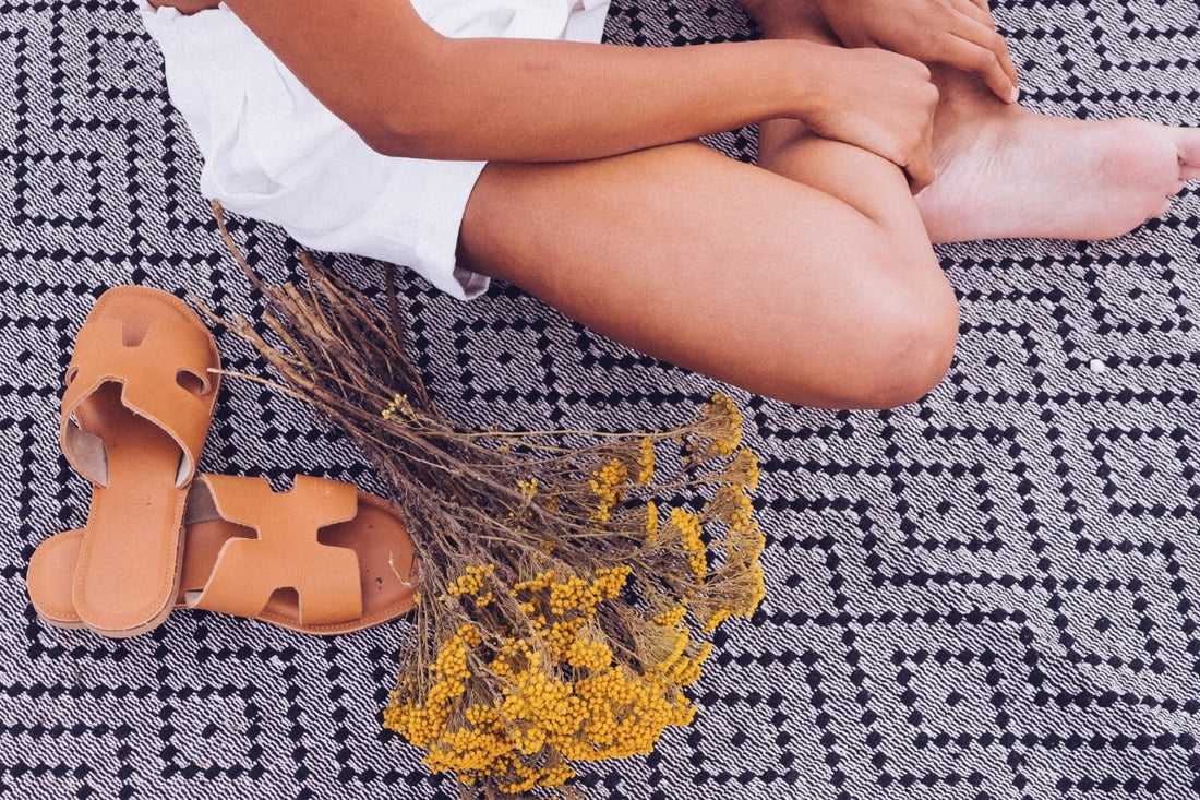 Woman sitting on black and white textured blanket Her legs are crossed and she is next to brown leather sandals and a bouquet of dried yellow flowers. Her face is not visible. She is wearing a white dress. Her skin in tanned.